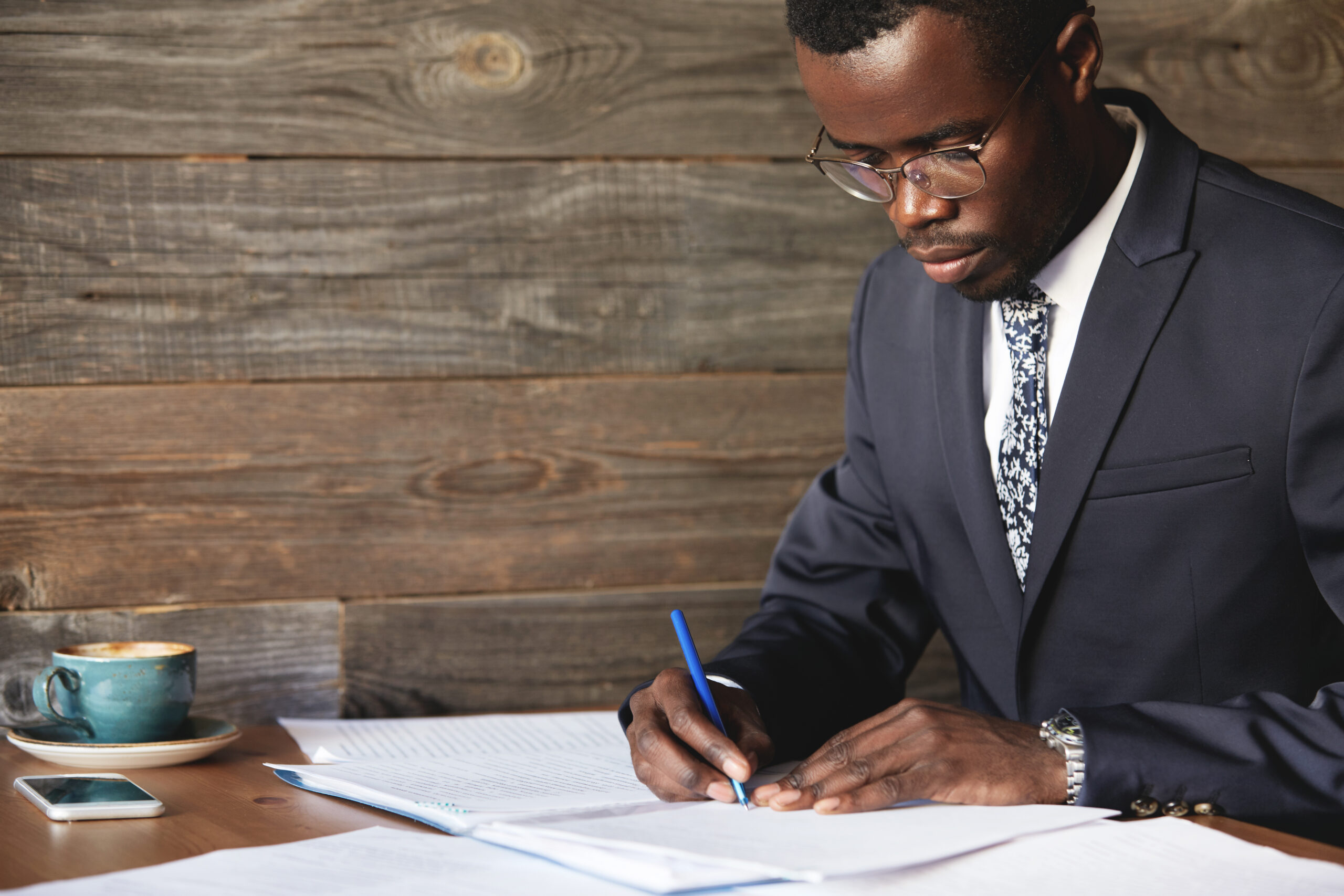 Serious black corporate worker in formal suit and spectacles signing a lucrative contract with concentrated expression while having morning cappuccino at a cafe, sitting against wooden wall background